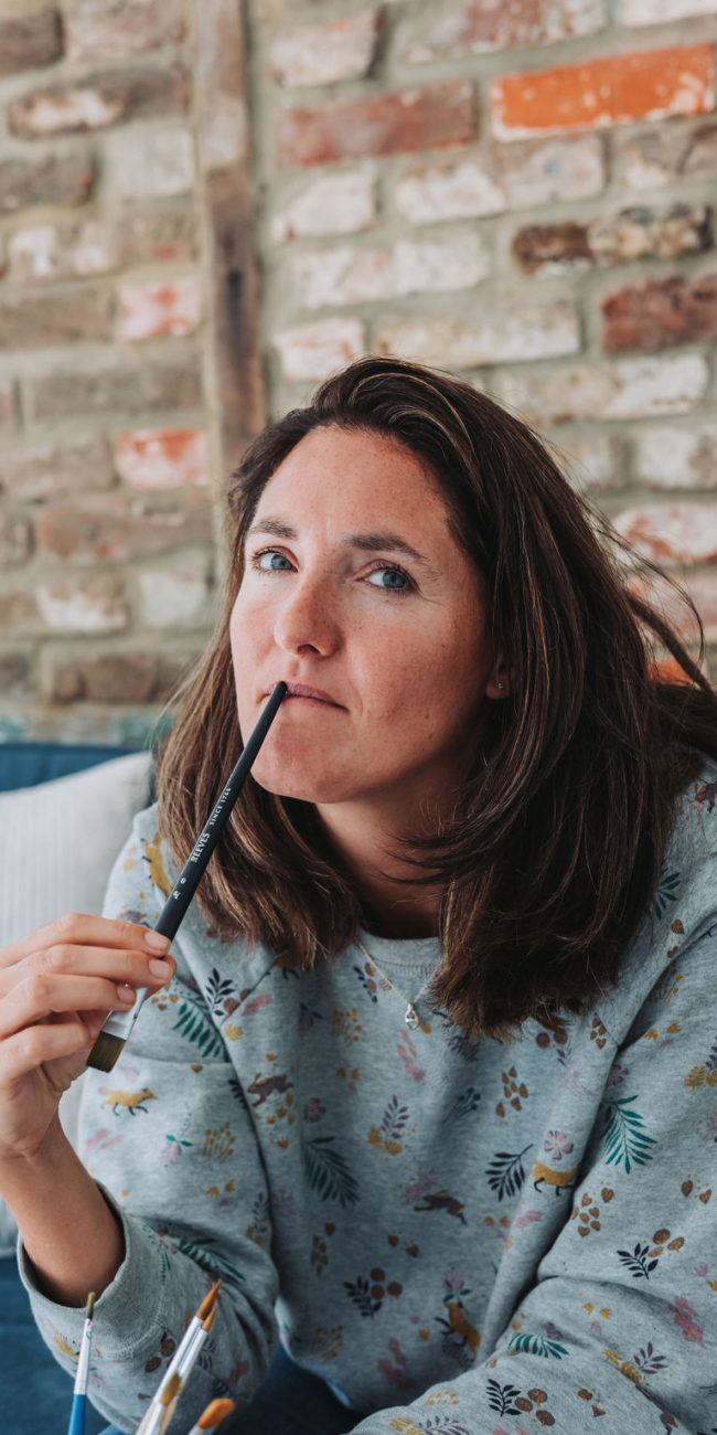 Katie holds a paintbrush, resting it near her mouth as she contemplates.